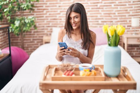 Téléchargez les photos : Jeune belle femme hispanique ayant petit déjeuner cadeau en utilisant smartphone dans la chambre - en image libre de droit