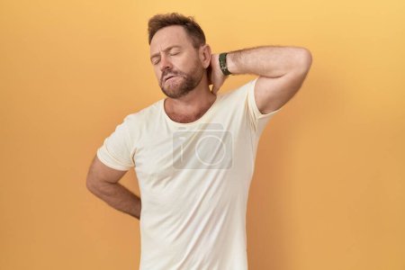 Foto de Middle age man with beard standing over yellow background suffering of neck ache injury, touching neck with hand, muscular pain - Imagen libre de derechos
