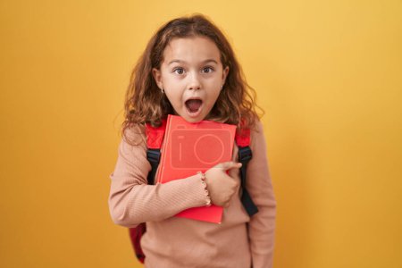 Foto de Pequeña chica caucásica usando mochila de estudiante y sosteniendo libro asustado y sorprendido con la boca abierta para sorpresa, cara de incredulidad - Imagen libre de derechos