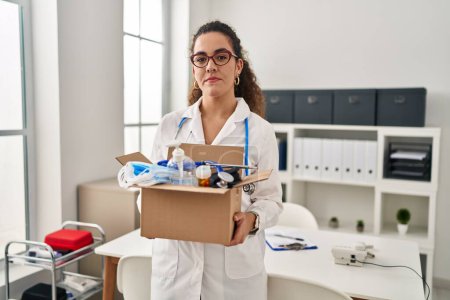 Foto de Young hispanic woman holding box with medical items relaxed with serious expression on face. simple and natural looking at the camera. - Imagen libre de derechos