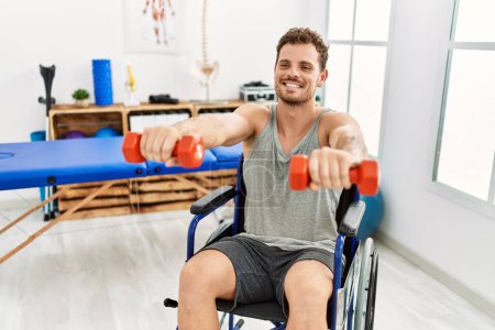 Photo for Young hispanic man doing rehab using dumbbells sitting on wheelchair at clinic - Royalty Free Image