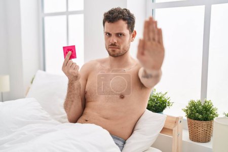 Téléchargez les photos : Young hispanic man holding condom in the bed with open hand doing stop sign with serious and confident expression, defense gesture - en image libre de droit