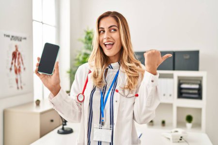 Téléchargez les photos : Jeune femme blonde médecin travaillant à la clinique montrant écran smartphone pointant pouce vers le côté souriant heureux avec bouche ouverte - en image libre de droit