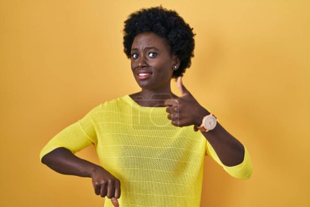 Téléchargez les photos : Jeune femme africaine debout au-dessus de studio jaune faisant pouces vers le haut et vers le bas, désaccord et expression d'accord. conflit fou - en image libre de droit