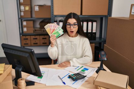 Foto de Young hispanic woman working at small business ecommerce holding money scared and amazed with open mouth for surprise, disbelief face - Imagen libre de derechos