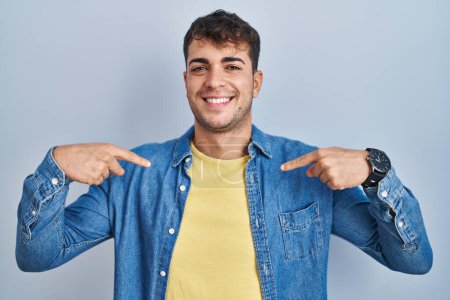 Foto de Young hispanic man standing over blue background looking confident with smile on face, pointing oneself with fingers proud and happy. - Imagen libre de derechos