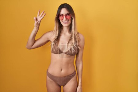 Foto de Mujer hispana joven vistiendo bikini sobre fondo amarillo mostrando y apuntando hacia arriba con los dedos número tres mientras sonríe confiada y feliz. - Imagen libre de derechos