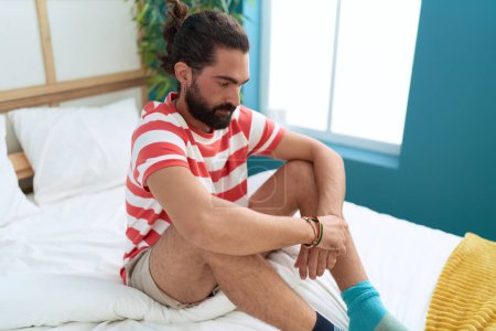 Téléchargez les photos : Young hispanic man sitting on bed with serious expression at bedroom - en image libre de droit