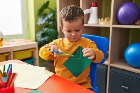 Téléchargez les photos : Adorable garçon caucasien coupant du papier assis sur la table à la maternelle - en image libre de droit
