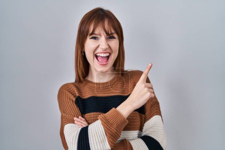 Foto de Young beautiful woman wearing striped sweater over isolated background with a big smile on face, pointing with hand finger to the side looking at the camera. - Imagen libre de derechos