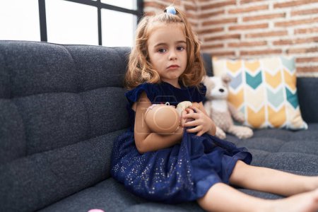 Photo for Adorable hispanic girl hugging baby doll sitting on sofa at home - Royalty Free Image