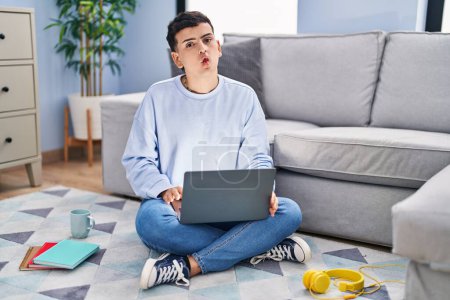 Photo for Non binary person studying using computer laptop sitting on the floor making fish face with lips, crazy and comical gesture. funny expression. - Royalty Free Image