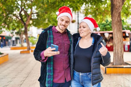 Foto de Mother and son wearing christmas hat using smartphone at park - Imagen libre de derechos