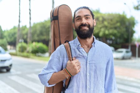 Téléchargez les photos : Jeune homme hispanique musicien souriant confiant tenant étui de guitare à la rue - en image libre de droit