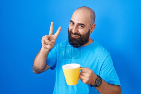 Foto de Joven hispano con barba y tatuajes bebiendo una taza de café sonriendo mirando a la cámara mostrando los dedos haciendo señal de victoria. número dos. - Imagen libre de derechos
