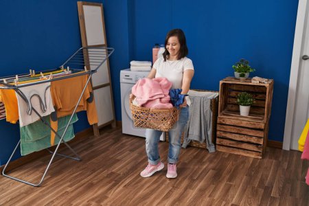 Foto de Síndrome de Down mujer sonriendo con confianza celebración cesta con ropa en la sala de lavandería - Imagen libre de derechos