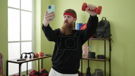 Foto de Young redhead man using dumbbells training and making selfie by smartphone at sport center - Imagen libre de derechos