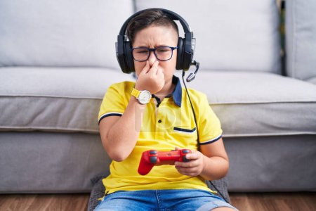 Foto de Young hispanic kid playing video game holding controller wearing headphones smelling something stinky and disgusting, intolerable smell, holding breath with fingers on nose. bad smell - Imagen libre de derechos