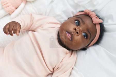 Téléchargez les photos : Bébé afro-américain couché sur le lit avec une expression détendue dans la chambre - en image libre de droit