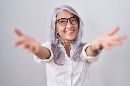Foto de Middle age woman with tattoos wearing glasses standing over white background looking at the camera smiling with open arms for hug. cheerful expression embracing happiness. - Imagen libre de derechos