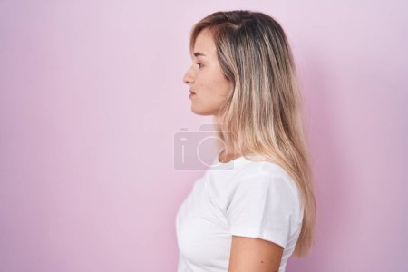 Foto de Mujer rubia joven de pie sobre fondo rosa mirando a un lado, pose de perfil relajante con cara natural y sonrisa confiada. - Imagen libre de derechos
