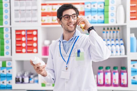 Foto de Joven farmacéutico hispano sosteniendo la botella de pastillas hablando en el teléfono inteligente en la farmacia - Imagen libre de derechos