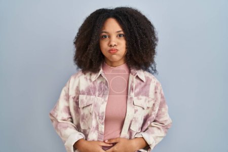 Foto de Young african american woman standing over blue background puffing cheeks with funny face. mouth inflated with air, crazy expression. - Imagen libre de derechos