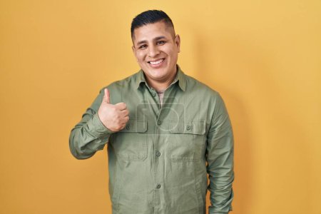 Foto de Hispanic young man standing over yellow background doing happy thumbs up gesture with hand. approving expression looking at the camera showing success. - Imagen libre de derechos