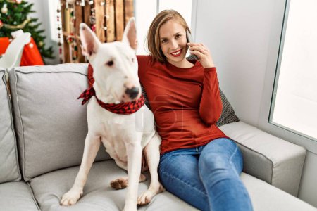 Téléchargez les photos : Jeune femme caucasienne parlant sur le smartphone assis avec chien par arbre de Noël à la maison - en image libre de droit