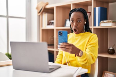 Téléchargez les photos : Belle femme noire faisant appel vidéo avec smartphone effrayé et étonné avec la bouche ouverte pour la surprise, visage incrédule - en image libre de droit