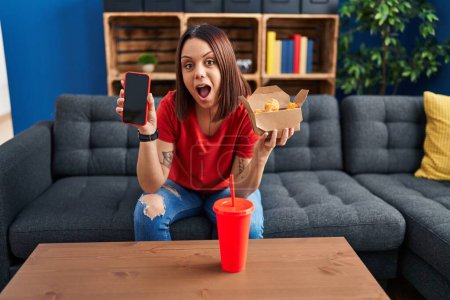 Foto de Young hispanic woman eating fast food showing smartphone screen afraid and shocked with surprise and amazed expression, fear and excited face. - Imagen libre de derechos