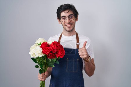 Téléchargez les photos : Young hispanic man holding bouquet of white and red roses doing happy thumbs up gesture with hand. approving expression looking at the camera showing success. - en image libre de droit