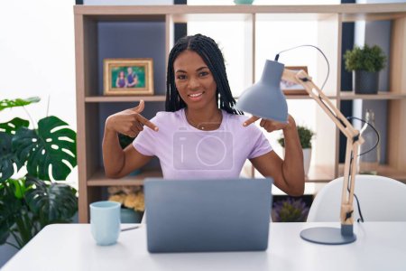 Foto de African american woman with braids using laptop at home looking confident with smile on face, pointing oneself with fingers proud and happy. - Imagen libre de derechos