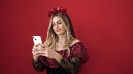 Foto de Mujer rubia joven usando teléfono inteligente que tiene fiesta de Halloween sobre fondo rojo aislado - Imagen libre de derechos