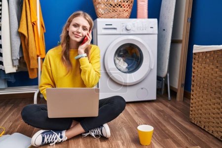 Téléchargez les photos : Jeune femme blonde utilisant un ordinateur portable parlant sur smartphone attendant la machine à laver à la buanderie - en image libre de droit