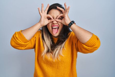 Photo for Young hispanic woman standing over isolated background doing ok gesture like binoculars sticking tongue out, eyes looking through fingers. crazy expression. - Royalty Free Image