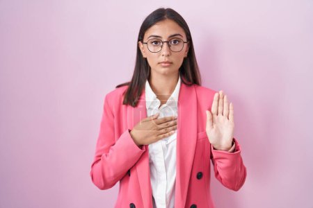 Foto de Young hispanic woman wearing business clothes and glasses swearing with hand on chest and open palm, making a loyalty promise oath - Imagen libre de derechos
