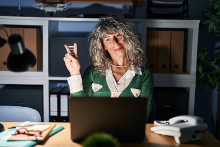 Téléchargez les photos : Femme d'âge moyen travaillant la nuit à l'aide d'un ordinateur portable souriant en regardant la caméra montrant les doigts faisant signe de victoire. numéro deux. - en image libre de droit