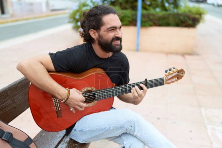 Téléchargez les photos : Jeune homme hispanique musicien jouant de la guitare classique assis sur un banc au parc - en image libre de droit