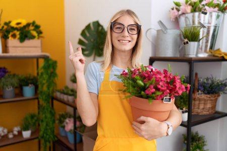 Foto de Young caucasian woman working at florist shop holding plant smiling happy pointing with hand and finger to the side - Imagen libre de derechos