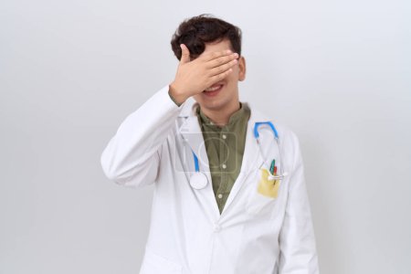 Téléchargez les photos : Young non binary man wearing doctor uniform and stethoscope smiling and laughing with hand on face covering eyes for surprise. blind concept. - en image libre de droit