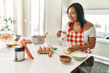 Téléchargez les photos : Hispanic brunette woman preparing cake looking at online recipe at the kitchen - en image libre de droit