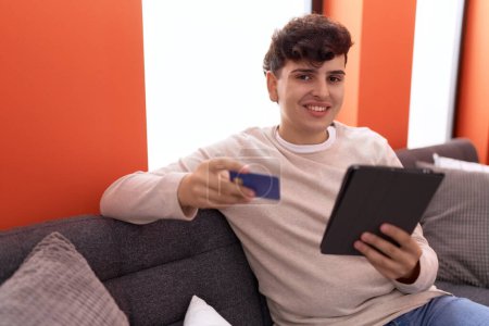 Téléchargez les photos : Homme non binaire utilisant le pavé tactile et la carte de crédit assis sur le canapé à la maison - en image libre de droit