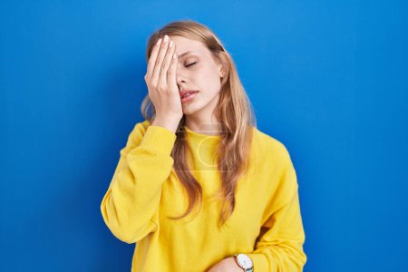 Photo for Young caucasian woman standing over blue background yawning tired covering half face, eye and mouth with hand. face hurts in pain. - Royalty Free Image