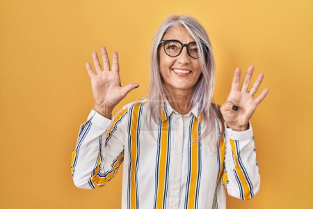 Foto de Mujer de mediana edad con el pelo gris de pie sobre el fondo amarillo con gafas que muestran y señalan hacia arriba con los dedos número nueve mientras sonríe confiado y feliz. - Imagen libre de derechos