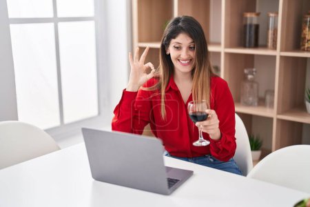 Photo for Young brunette woman doing video call drinking red wine doing ok sign with fingers, smiling friendly gesturing excellent symbol - Royalty Free Image
