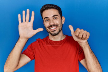 Téléchargez les photos : Young hispanic man with beard wearing red t shirt over blue background showing and pointing up with fingers number six while smiling confident and happy. - en image libre de droit