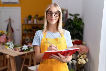Téléchargez les photos : Jeune femme blonde fleuriste souriant écriture confiante sur ordinateur portable à la boutique de fleurs - en image libre de droit