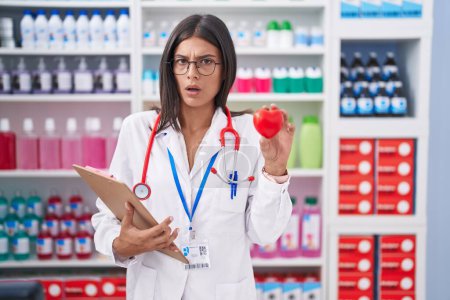 Téléchargez les photos : Brunette young woman working at pharmacy drugstore holding red heart in shock face, looking skeptical and sarcastic, surprised with open mouth - en image libre de droit