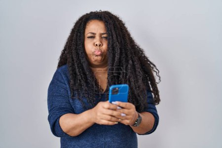 Téléchargez les photos : Plus size hispanic woman using smartphone typing message puffing cheeks with funny face. mouth inflated with air, catching air. - en image libre de droit